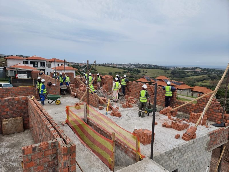 A bricklaying team building walls on a completed slab with coastal hills behind.