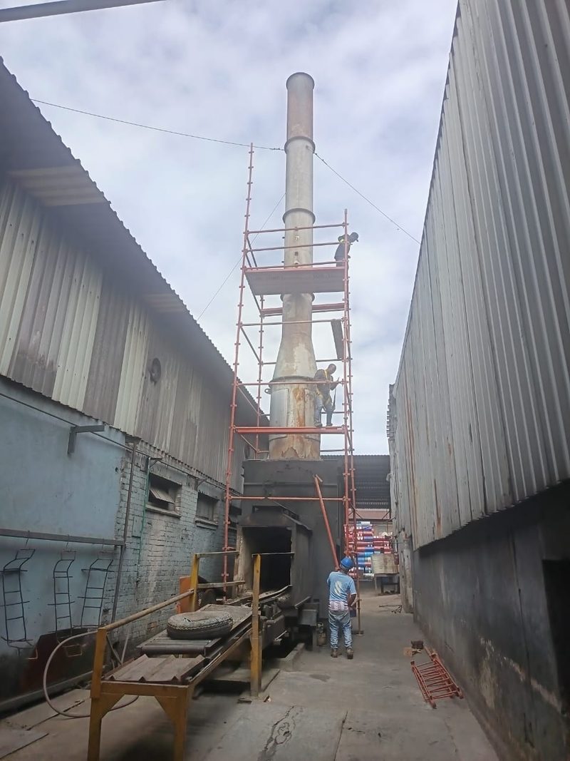 Scaffolding erected around a tall industrial flue between two buildings.